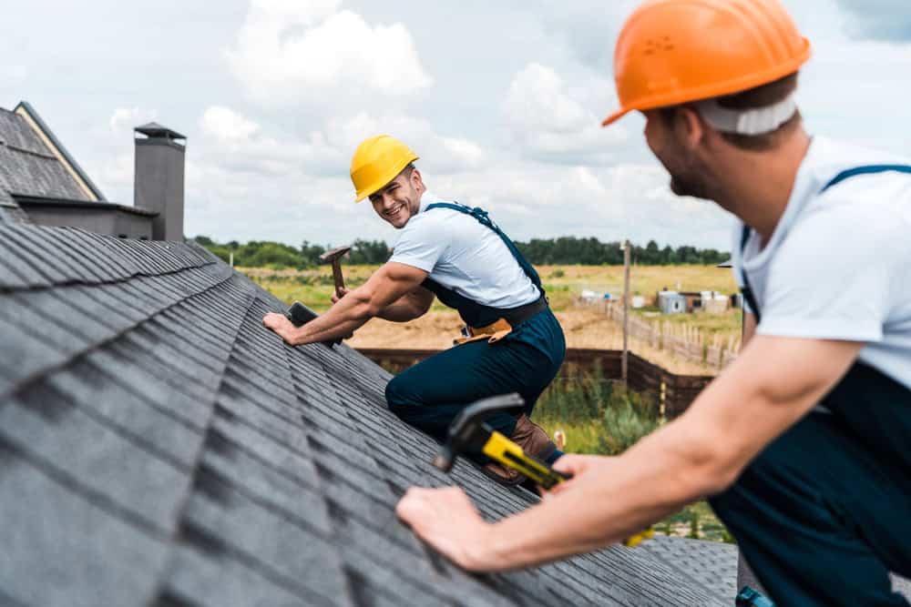 Two construction workers in white shirts and orange safety helmets are working on a sloped roof. One is smiling and holding a hammer, while the other is crouched with a tool in hand. The sky is partly cloudy in the background.