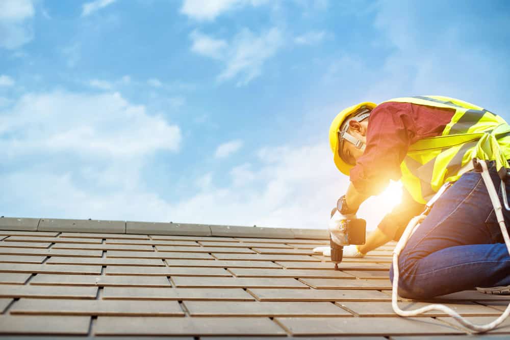 A construction worker in a hard hat and safety vest kneels on a roof, using a power tool. The sky is clear with a bright sun in the background.