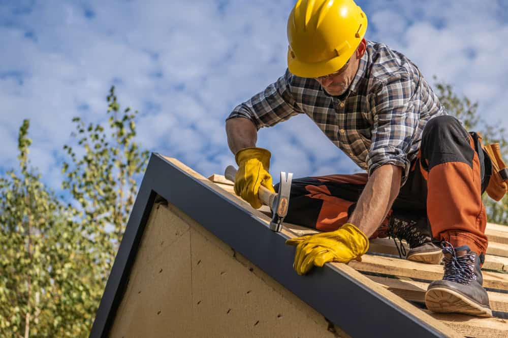 A construction worker in a yellow hard hat and gloves hammers nails into the roof edge of a wooden structure. He's wearing a plaid shirt and orange pants. The sky is partly cloudy, and trees are visible in the background.